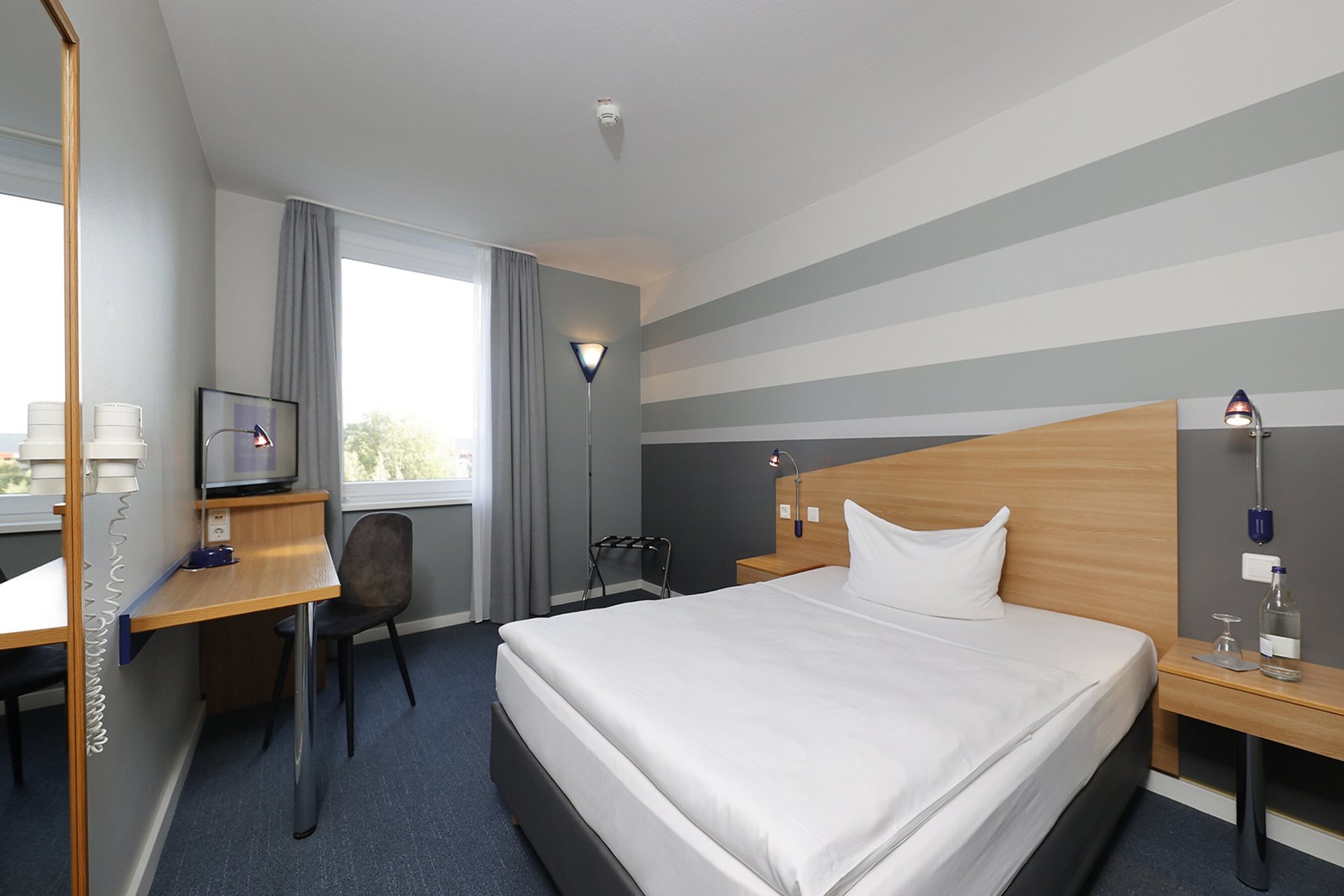 Unser Hotel an der Messe: die Bildergalerie Modernes Hotelzimmer mit großem Bett, Schreibtisch und Fenster mit grauen Vorhängen